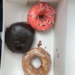 Sour Cream Cake Donut - Three donuts with various toppings are placed inside a white box, including a pink frosted donut with sprinkles, a chocolate glazed donut, and a glazed donut with a bite taken out.