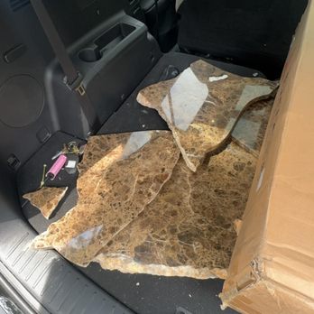 Arch Moving - A large marble slab with a brown and beige pattern is positioned in the trunk of a vehicle, covering the trunk floor and extending onto the back seat area. The vehicle's interior is visible, including the back of the front seats and the side panel. A cardboard box is placed next to the marble slab, and a set of keys and a lighter are on the trunk floor. The photo appears to be taken during daytime, with some outdoor elements visible through the window. - Movers Near Me - Saint Ann, Missouri