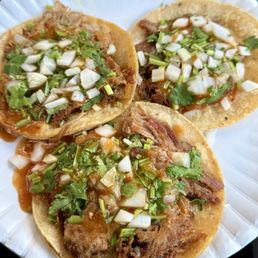Carnitas Tacos - Three carnitas tacos with chopped onions and cilantro are served on a white disposable plate, garnished with sauce.