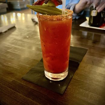 Ray's Corner Cafe - Bloody Mary - A tall glass of Bloody Mary cocktail with a salted rim is placed on a dark wooden table. The drink contains tomato juice and is garnished with a pickle spear, a strip of bacon, and a piece of tomato and lettuce. The background shows a person in a plaid shirt and a bottle of alcohol, indicating a restaurant or bar environment. - Salad Near Me - Montrose, Colorado