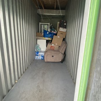 Extra Space Storage - The image shows a small storage unit with metal walls and a concrete floor. Inside, there are stacked cardboard boxes, a beige recliner chair, and miscellaneous household items including bottled water, a small white cabinet, and bags. The ceiling has exposed wooden beams and a metal duct. The space appears cluttered and used for storage. - Self Storage Near Me - Boca Raton, Florida