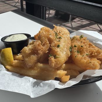 Rooted Craft Kitchen - Fish and Chips - A serving of fried fish fillets with French fries, accompanied by tartar sauce and a lemon wedge, on a white table outdoors. - New American Near Me - Boulder, Colorado