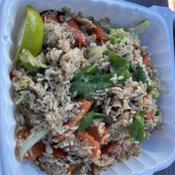 Thai Time - Thai Fried Rice - A white plastic takeout container holds Thai fried rice with mixed vegetables such as carrots, cabbage, and possibly mushrooms. The dish is garnished with fresh cilantro and a lime wedge, and is placed on a dark outdoor surface. The rice appears well-cooked with a variety of textures and colors, typical of Thai cuisine. - Food Trucks Near Me - Austin, Texas