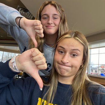 Culver's - unhappy customers - Two young women are in an indoor setting with large windows behind them, allowing natural light to illuminate their faces. The woman in the foreground wears a navy blue sweatshirt with yellow lettering and has long, light brown hair. The woman behind her has wavy, light brown hair and wears a gray sweatshirt. Both are making thumbs down gestures, with the woman in the foreground displaying a neutral or slightly amused expression, and the woman behind smiling subtly. - Ice Cream & Frozen Yogurt Near Me - Lafayette, Indiana