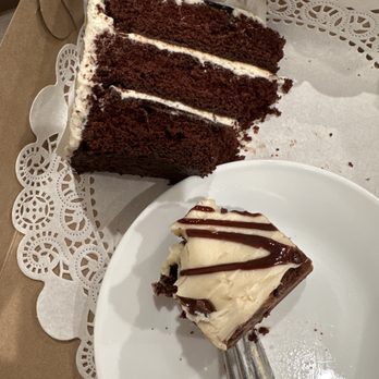 The Chocolate - A slice of chocolate cake with white frosting and a slice of cheesecake with chocolate drizzle are displayed on a white plate and in a bakery box. The chocolate cake has three layers with frosting between each layer, and the cheesecake has a chocolate crust with a creamy top. The image captures the desserts in a casual setting, likely after purchase from a bakery or cafe. - Desserts Near Me - Orem, Utah
