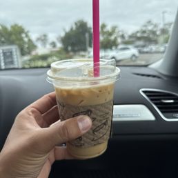 Banana Bread Latte - A hand is holding a cold coffee beverage in a clear plastic cup with a pink straw, inside a car. The cup has a cardboard sleeve, and the background shows a parking lot with cars and trees under a cloudy sky.