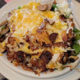 A breakfast or brunch dish featuring melted cheese, bacon, mushrooms, and vegetables in a white bowl, with a pancake visible in the background.