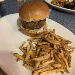 Hamburger and fries - A hamburger with a beef patty, lettuce, and bun is served on a white plate. The burger includes a toasted bun, a beef patty, and some lettuce. A portion of French fries is placed next to the burger. In the background, there is a glass of beer and another plate with food, indicating a dining setting at a restaurant.