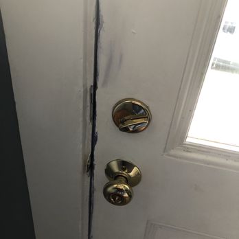 First Choice Masonry - Sloppy paint job got on inside of the door - A white door features a vertical crack along its edge, with the lock mechanism exposed and missing its cover. The door has a window on the right side, allowing light to pass through. The door frame shows signs of damage or wear near the crack. - Roofing Near Me