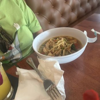 The Joint - Chicken Veggie soup of the day. - A bowl of chicken veggie soup with noodles and vegetables is on a wooden table, with a fork resting on a napkin nearby. A glass of water and a yellow drink with a lemon slice are also on the table. Part of a person wearing a green shirt with sunglasses hanging from the collar is visible in the image. The setting appears to be a casual dining environment, possibly a restaurant or cafe. - Beer, Wine & Spirits Near Me - Boston, Massachusetts
