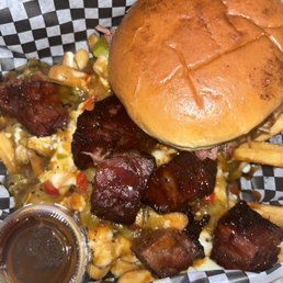 a burger and fries - A barbecue meal includes smoked burnt ends with a glossy, caramelized exterior, placed on a checkered paper-lined tray. A sandwich bun is positioned on top of the burnt ends, with French fries visible underneath. A small plastic container of barbecue sauce is included in the tray. The food appears freshly prepared, with the burnt ends showing a dark, flavorful crust and tender interior.