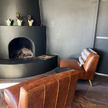 El Verde Inn - Lounge area - Two brown leather armchairs are arranged near a black fireplace with a curved opening, set against dark gray walls. The chairs are placed on a brown animal hide rug. One chair has a striped blanket draped over its back. The fireplace has three small potted plants on top, each in a different colored pot. A door with a metallic handle is visible on the right side of the image. - Hotels Near Me - Albuquerque, New Mexico