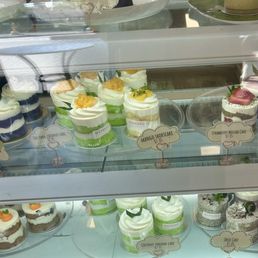 A bakery display case contains multiple small cakes and pastries, including coconut pandan cake, mango shortcake, strawberry matcha cake, and Oreo cake. Each item is labeled with a small sign indicating its name and price. The cakes are decorated with cream, fruit, and other toppings, arranged on clear trays inside the case. The background shows a reflection of the bakery interior and some additional baked goods on the upper shelf.