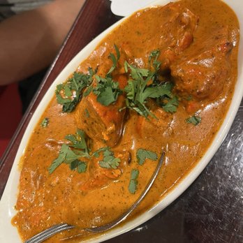 Namaste - A bowl of Indian chicken curry features a thick, orange-colored sauce with visible herbs and spices. The dish contains pieces of chicken and is garnished with fresh cilantro leaves. It is served in a white oval bowl placed on a dark wooden table, with a spoon resting inside the bowl. The background shows part of a person's arm and a white plate. - Indian Near Me - Rio Rancho, New Mexico