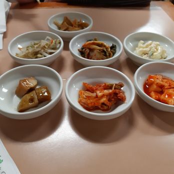 Babo Jumak - A variety of Korean side dishes in small white bowls on a table, with a person using chopsticks in the background. - Korean Near Me - Mesa, Arizona