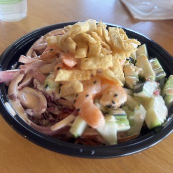 Toss’d - Bang Bang Shrimp - A seafood salad with shrimp, chopped cucumbers, and vegetables, coated in a creamy dressing, topped with fried onions, served in a black plastic bowl on a wooden table. In the background, there is a partially visible drink and a red-and-white checkered container. - Wraps Near Me - Temecula, California