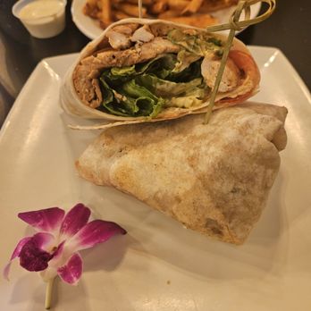 Mimosas cafe - A chicken wrap with lettuce, chicken slices, and other fillings is presented on a white plate, secured with a skewer. The wrap is accompanied by a side of French fries and a small container of dipping sauce. A pink and white flower garnish is placed on the plate, and the background shows a table setting with additional condiments and food items. - Breakfast & Brunch Near Me - Merced, California