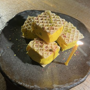 Rumis Kitchen - Four layered dessert squares with a yellow exterior and white top, garnished with crushed pistachios and pink flower petals, on a dark ceramic plate with a lit birthday candle. - Persian/Iranian Near Me - Houston, Texas