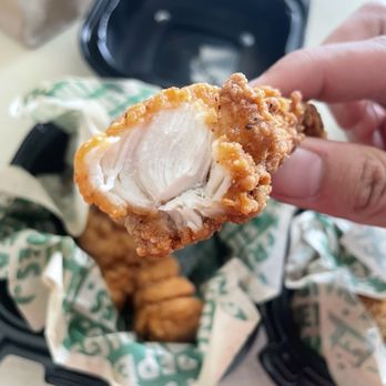 Wingstop - A person holds a piece of fried chicken, showing the white meat inside, above a black takeout container filled with more fried chicken pieces. The container is lined with branded paper, and the background includes a blurred table surface and a paper bag. The image emphasizes the crispy exterior and tender interior of the fried chicken. - Chicken Wings Near Me - Albany, New York