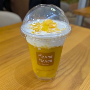 Mango Mango Dessert - Mango Coconut Smoothie-O - A mango coconut smoothie is served in a clear plastic cup with a domed lid, topped with pieces of fruit and whipped cream. The cup has a Mango Mango Dessert logo and is placed on a wooden table. The background shows blurred figures and a casual setting. - Desserts Near Me - Plainview, New York