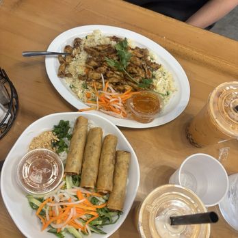 Wrap N Roll - The image shows two white plates of Vietnamese food on a wooden table. One plate contains four spring rolls with a side of chopped vegetables, peanuts, and a small container of dipping sauce. The other plate has grilled meat served over rice, garnished with herbs, with shredded carrots and cucumbers on the side. There are three bubble tea drinks with straws and plastic lids, along with empty plastic cups. A person with tattoos on their arm is partially visible in the background. - Vietnamese Near Me - Albuquerque, New Mexico