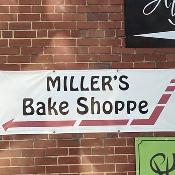 Miller's Bake Shoppe - Street Sign - A white fabric banner with black text reading 'MILLER'S Bake Shoppe' and a red arrow pointing left is displayed against a brick wall. - Bakeries Near Me - Stuarts Draft, Virginia