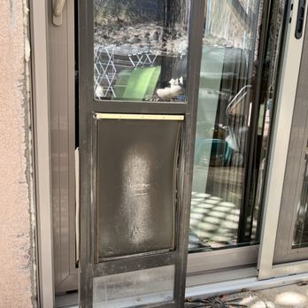 Flawless Results - Original dog door next to new patio door, which is too short. - The image shows a metal pet door installed in a sliding glass door frame, with a screen door above it. The pet door has a transparent flap and is positioned at the bottom of the screen door. The surrounding area includes a concrete patio with some shadows and outdoor furniture visible through the glass. The installation appears to be part of a home improvement or door replacement project. - Windows Installation Near Me - Albuquerque, New Mexico