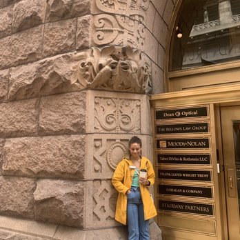 Inside Chicago Walking Tours - Rookery building & Kylie, our wonderful guide. - A woman with dark hair tied back, dressed in a yellow jacket, turquoise top, and jeans, stands on a sidewalk next to a building with ornate stone carvings and a decorative stone relief. She holds a smartphone in her right hand and is smiling. The building features detailed stonework, including carved faces and decorative patterns, and a sign listing various businesses such as Dean Optical and Moody-Nolan. The scene appears to be in an urban area with reflections of other buildings in the glass above the sign. - Architectural Tours Near Me - Chicago, Illinois
