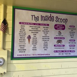 List of flavors - The menu board for The Inside Scoop ice cream shop lists various flavors and options, including homemade ice cream, yogurt, sorbet, and smoothies. It features sections for different types of treats such as ice cream cakes and no-sugar-added options. The board is framed in green and purple, with a small image of an ice cream cone, and is mounted on a yellow wall. An American flag is visible to the left of the board, and a utility meter is mounted below it.