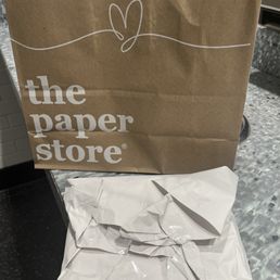 Rush no care job! - A brown paper shopping bag from The Paper Store features white printed text and a simple line drawing of a heart with a line extending from it. The bag has twisted paper handles and is positioned on a gray speckled countertop, with a crumpled white paper-wrapped package in front of it. The background includes a tiled wall and part of a metallic surface.