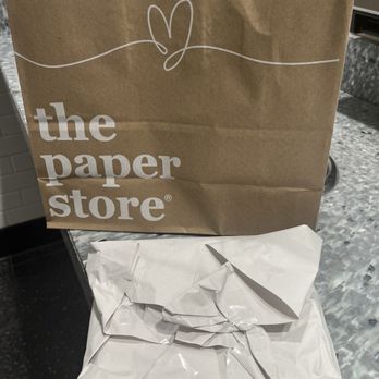 The Paper Store - Rush no care job! - A brown paper shopping bag from The Paper Store features white printed text and a simple line drawing of a heart with a line extending from it. The bag has twisted paper handles and is positioned on a gray speckled countertop, with a crumpled white paper-wrapped package in front of it. The background includes a tiled wall and part of a metallic surface. - Gift Shops Near Me - Cranston, Rhode Island