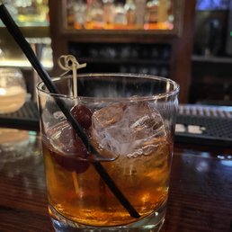 Old fashioned - A glass of cocktail with ice, cherries, and a black straw on a wooden bar counter, with a blurred bar background.