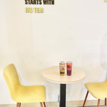 ITS TEA - Interior - Inside a tea shop, a small round table holds two cups of tea or juice, with two yellow chairs on either side. A sign on the wall promotes tea. - Bubble Tea Near Me - Toronto, Ontario
