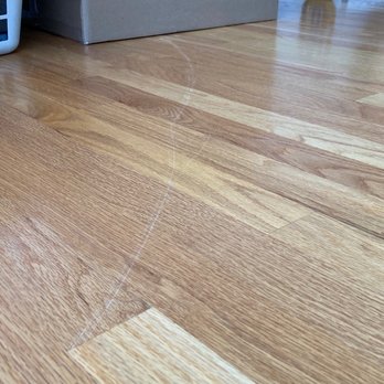 BZ Movers - A photograph captures a section of hardwood flooring with a prominent crack or scratch extending across the surface. The floorboards are light to medium wood tones, and the crack appears to be a surface imperfection. In the background, a laundry basket and a cardboard box are partially visible, indicating an indoor setting. - Movers Near Me - Long Grove, Illinois