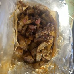 Madre's Breakfast Taco Make It Your Way! - A breakfast taco contains cooked chorizo, scrambled egg, melted cheese, and diced potato filling, wrapped in a soft tortilla. The taco is placed on aluminum foil, with some of the filling visible and spilling slightly from the tortilla. The image shows a close-up view of the taco, highlighting the ingredients inside.