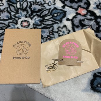 Elevation Yarn - A cardboard box and a paper bag with Elevation Yarn & Co logos are displayed on a floral-patterned rug, with metal clips attached to the paper bag. - Knitting Supplies Near Me - Littleton, Colorado