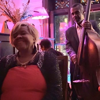 Brownstone JAZZ - A woman with short blonde hair, wearing a black jacket with embellishments and a red top, is smiling in a jazz venue. A man in a suit is playing a double bass nearby. The venue has colorful lighting, large windows, and decorative elements like lamps and plants, creating a lively atmosphere. - Jazz & Blues Near Me - Brooklyn, New York