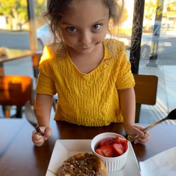 CB Kitchen - A girl with curly hair and blue eyes, wearing a yellow dress, stands at a cafe table with a pastry and a bowl of strawberries. - Burgers Near Me - Willows, California
