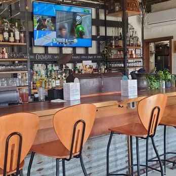 Crow and Cart - Bar area - A bar with a row of wooden chairs, a TV screen showing a sports game, and a menu board on the wall in a restaurant setting. - Cocktail Bars Near Me - Eugene, Oregon