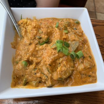 Masala - Navrattan Korma - A square white bowl holds Navrattan Korma, an Indian vegetable curry featuring a thick, orange-colored sauce with visible pieces of vegetables and spices. The dish is garnished with chopped green onions on top. The bowl is placed on a wooden table, and part of a person's arm and clothing are visible in the background, indicating a restaurant setting. - Indian Near Me - Somerville, Massachusetts