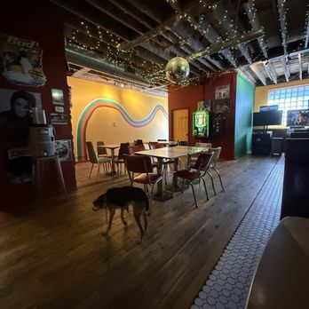 Circular Lounge - Nice open space - The photo depicts the interior of a lounge or bar with a dark ceiling decorated with string lights and a disco ball. The walls are painted in vibrant colors, including a yellow wall with a rainbow-like mural and a red wall with a door. There are tables and chairs arranged in the space, and a vintage jukebox is visible near the red wall. A dog is walking across the wooden floor, which has a section of white hexagonal tiles along the edge. The space appears cozy and retro-themed, with a mix of lighting and decor elements. - Lounges Near Me - Denver, Colorado