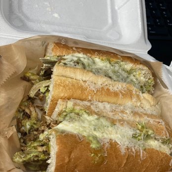 Mina Bistro - A sandwich with melted cheese, shredded cheese, and vegetables such as lettuce and possibly mushrooms is placed inside a foam takeout container. The sandwich appears to be made with a soft bread roll, and the filling is visible from the side. The container is lined with paper, and the background shows part of a keyboard and a surface, indicating it might be in an office or work setting. - Venezuelan Near Me - Miami, Florida