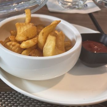The Kitchen - A white ceramic bowl contains French fries, placed on a white plate on a restaurant table. Next to the bowl is a small black container of ketchup. The table surface is wooden, with some glassware and other items partially visible in the background. - Bars Near Me - Carmel, Indiana