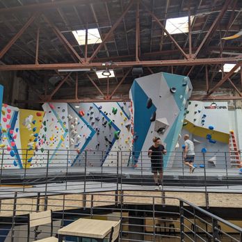 The Oaks - Upstairs slab and beginner climbing area - The image shows an indoor bouldering gym with multiple climbing walls of different colors and angles, equipped with holds for climbing. Several people are actively climbing or resting on the padded floor area. The gym has an industrial ceiling with exposed beams and skylights, and a metal railing surrounds the climbing area. There are tables and chairs in the foreground, indicating a space for resting or socializing. - Rock Climbing Near Me - Berkeley, California
