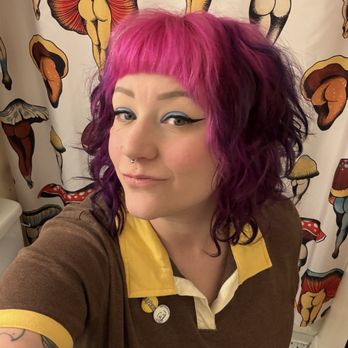 Rockit Hair Studio - A woman with pink and purple curly hair, styled with a blunt fringe, takes a selfie in a room with a mushroom-themed curtain featuring various colorful mushroom illustrations. She has makeup with winged eyeliner and blush, and a septum piercing. She wears a brown shirt with yellow accents and a white collar, and has tattoos on her arm. The setting appears to be a bathroom or dressing area with a tiled floor. - Hair Salons Near Me - Albuquerque, New Mexico