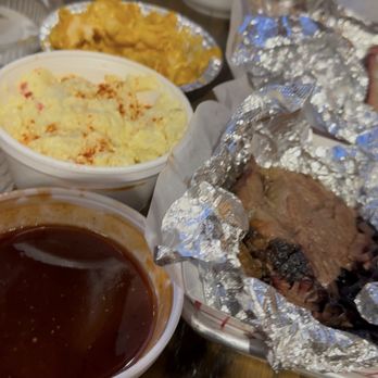 Test Kitchen BBQ - Brisket, potato salad, and baked Mac and cheese - The image shows a table with multiple food items including a baked potato topped with cheese, macaroni and cheese, and barbecue meat wrapped in aluminum foil. There are also bowls of barbecue sauces and a small container of what appears to be a side dish. The setting suggests a casual dining environment, possibly at a barbecue restaurant or gathering. - Barbeque Near Me - Waukegan, Illinois