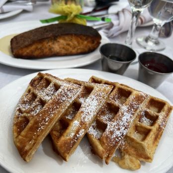 Patrick Steakhouse - Four waffles dusted with powdered sugar are arranged on a white oval plate on a restaurant table. In the background, there is a salmon fillet on a separate plate, along with two small containers of syrup, glasses of water, and champagne flutes. The table is set with utensils, napkins, and a decorative centerpiece, indicating a dining setting. - Steakhouses Near Me - Brooklyn, New York