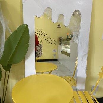 Ten Cake Studio - mirror and seatssz - The image shows a seating area with yellow tables and chairs positioned against a yellow wall. A decorative mirror with a white, irregular frame resembling dripping paint or slime is mounted on the wall, reflecting part of the room. The mirror is decorated with spider web and Halloween-themed decorations, including small hanging spiders and a pumpkin figure. The background includes a display case and some Halloween-themed wall decals, creating a festive atmosphere. - Patisserie/Cake Shop Near Me - Brooklyn, New York