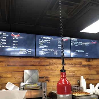 Bartley's Bar-B-Que - Menu - Two digital menu screens mounted on a wooden wall display food options including barbecue plates, sandwiches, and value meals. The left screen lists various meat plates with prices, while the right screen features sandwich options, baked potato choices, and frozen ribs. Below the screens, a red lamp, cups, and food service equipment are visible, indicating a casual dining or takeout restaurant setting. - Barbeque Near Me - Grapevine, Texas