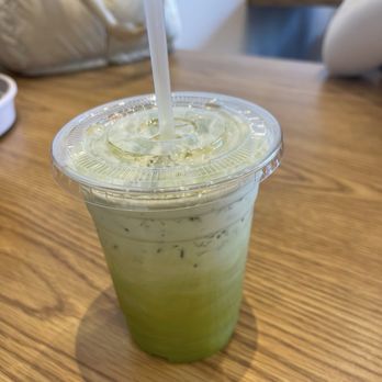 Pho Anan - Green Thai Tea - A beverage with green and white layers in a clear plastic cup, topped with a plastic lid and straw, placed on a wooden table. - Vietnamese Near Me - Glendale, Arizona